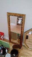 Rectangular bevelled mirror with a wooden frame leaning against a wall, showing textured brown frame and clear reflective glass.