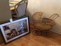 Photo showing the framed print of Raphael's Little Angels leaning against a chair and a vintage wicker pram with large wheels beside it on wooden floor.
