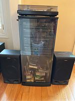 Full view of Sony stereo system with cabinet and speakers, showing stereo components stacked inside black glass door cabinet, two black Sony speakers on sides, and various DVDs/CDs in cabinet.