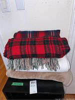 Stack of five folded throw blankets including red plaid, beige/tan, white, and dark-colored plaid blankets.