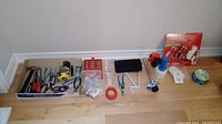 Full lot of assorted hand tools and electrical items, laid out on a wooden floor showing two Snap-on tape measures, electrical tape, metal bracket, power outlet plates, a small flashlight, scotch tape, and other items.