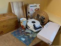 Photo showing kitchen corner countertop with a wooden flour box labeled 'Fleurs', Phillips Belgium waffle maker with its box, Phillips food saver, two wooden cutting boards, a ceramic mug, woven basket with silverware, and two kitchen mats with cartoon chef prints.
