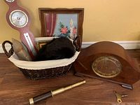 Overall lot on floor showing the woven basket with barometer, muskrat hat, framed needlepoint behind, Tasco brass monocular in front, Bertmar mantel clock and winding key on the right side.