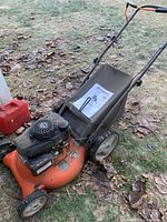 Side view of Husqvarna lawn mower 7021P with black Honda engine, orange base, black grass collection bag and attached manual on top, placed outside on grass and fallen leaves.