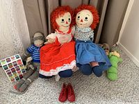 Full lot showing two Raggedy Ann dolls seated next to classic sock monkey, green plush caterpillar, and Campbell Soup box on carpeted floor.