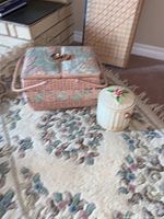 Photo of vintage wicker sewing basket with pastel floral fabric lids, cream ceramic jar with floral lid, and wool rug with floral pattern underneath.