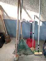 Wide shot of various garden tools including rakes, shovels, and brooms all with wooden handles, showing varying colors and types of heads and bristles.