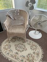 View of white wicker chair, two pedestal fans and oval floral wool rug.