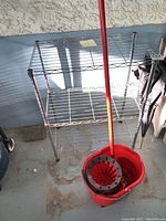 Metal wire utility stand with three shelves and red mop bucket with wringer attachment placed in front