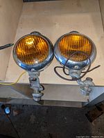 Pair of vintage round amber automotive lights with chrome housings and mounting brackets viewed from the front.