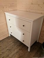 Photo of a white wooden dresser with three drawers against a wooden paneled wall on a wooden floor.