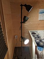 Full view of black 3 light corner lamp standing lamp with three adjustable shades, plugged into wall outlet next to wood-paneled wall and sofa.