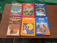 Photo of 6 vintage Atari 2600 game cartridges in original boxes arranged on a wooden surface. Games included are River Raid, Combat, Space Invaders, Haunted House, Pac-Man, and Defender. Boxes show wear and weathering.