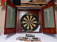 Open wooden dart board cabinet showing the Winmau Blade dart board inside with chalk scoreboards on cabinet doors and new dart sets on surface below