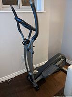 Side view of the elliptical exercise machine showing the foot pedals and handlebar structure against a wall and floor background.