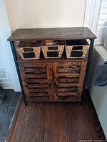Front view of wooden entrance hutch with three woven baskets on top placed against wall on hardwood floor.
