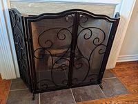Front view of the three-panel black metal decorative fireplace screen with intricate scroll-work and mesh backing installed in fireplace opening.