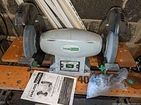 Front view of Hauss Mann bench grinder with both grinding wheels, adjustable rests, safety guards, power switch and manual on the bench.