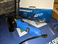 Blue MasterCraft 4.5 inch angle grinder with black protective guard and black detachable side handle placed in front of the original blue and white product box.