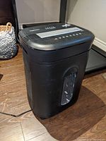 Full view of black Amazon Basics office paper shredder on wooden floor with visible pull-out bin and viewing window.
