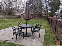 Full view of patio set showing round black metal table with four matching chairs and closed brown umbrella set up on stone patio.