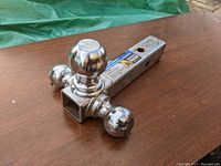 Photo of a chrome-finished trailer hitch with three different sized hitch balls mounted on the same hitch shank, placed on a wooden surface.