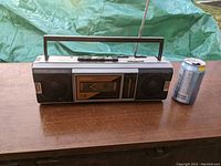 Front view of vintage portable boombox on wood surface next to soda can for size comparison, showing dual speakers, cassette compartment and antenna.