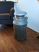 Full front view of the silver-colored metal milk can sitting on a wood floor with a dark sofa in the background.