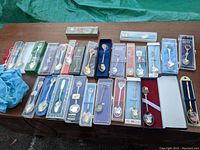 Group photo showing approximately 25 vintage souvenir spoons in original boxes and plastic packaging displayed on table