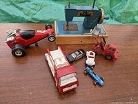 Photo showing seven vintage toys including a blue and red toy sewing machine on a wooden box, a red large fire truck, two miniature cars black and white, a long red race car, a blue single-seater race car, and a small red loader crane.