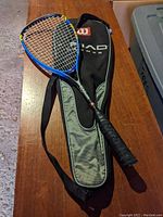 Wilson tennis racket with black and yellow strings, blue frame, and black grip next to black and gray Wilson carrying case on wooden table.