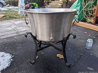 Large stainless steel bowl with drainage spout on black metal stand shown outside on pavement with surrounding outdoor items visible.