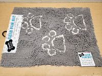 Top view of gray rectangular dog mat with white paw prints, showing label 'Comfy Pooch Clean Paw Mat' attached.