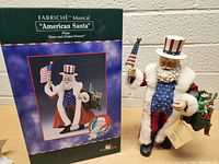 Photo of the Kurt Adler FABRICHE American Santa musical figurine and its box, showing the full figure, colors, and the flag it holds.