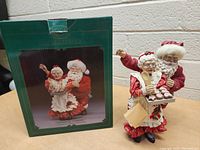 Photo of the full figurine showing Santa standing behind Mrs. Claus holding baking items and cookies, with the box in background.