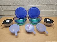 Photo showing 5 glass bowls with blue lids and 5 plastic bowls with translucent blue lids and handles on a wooden surface against a brick wall.