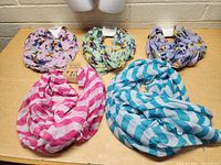 Five infinity scarves laid out on wooden table showcasing variety of patterns and colors including floral bird prints and stripes.