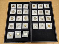 Two black trays containing 26 assorted foreign coins mounted in individual white holders with notes on country, denomination, and year.