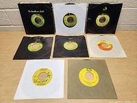 Complete view of eight Beatles 45 RPM records arranged on a table with various sleeves, mostly Apple Records. Some sleeves show wear and tears.