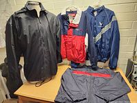 Photo showing four men’s jackets on display including black zip-up, navy and red jacket, navy blue hooded jacket, and grey jacket with red accents.
