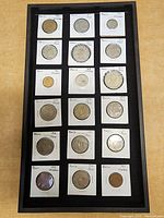 Top view of a black framed coin holder displaying Mexican coins from the 1950s to 1970s, each coin individually placed in a labeled cardboard holder.