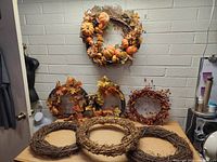 Group photo showing two decorative wreaths with artificial pumpkins and leaves, two wreaths with artificial leaves and berries, one wreath with purely berries, and three plain grapevine wreath bases.