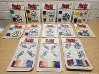 Photo of full lot of 14 vintage Makeit & Bakeit stained glass kits on a table showing a variety of designs and colored plastic cooking crystals in packaging.
