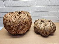 Two woven decorative pumpkins, one large and one smaller, shown side by side against a white brick wall and beige surface.