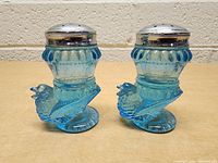 Pair of blue glass salt and pepper shakers shaped like chickens with metal tops on wooden surface.