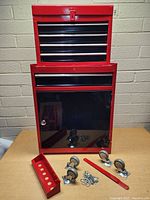 Front view of red tool chest cabinet with multiple drawers, lockable lower door, and four caster wheels and mounting hardware laid on table, also includes a red metal tool organizer tray.
