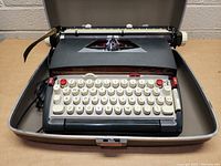 Front view of a vintage Sears typewriter seated inside its open gray plastic hard case, keys white with some red keys, ribbon visible, tested working.