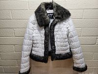 Front view of white Sugarfly puffy jacket with gray faux fur collar, cuffs, and hem hung on hanger against brick wall.