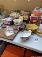 Full view of lot showing Crowning Touch cookware boxes, serving bowls, cheese plate with dome, fondue set box, and large pan on white table.
