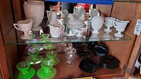 Shelf display of white milk glass and hobnail pitchers, bowls, hen on nest dish, footed egg cup, green uranium glass teacups and saucers, blackware plates and napkin holder.
